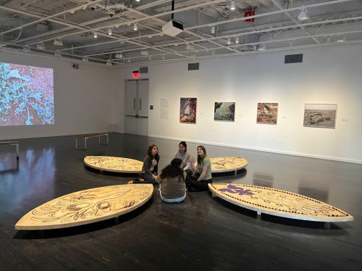 Four people sit on a circular wooden installation in an art gallery with displayed artworks.